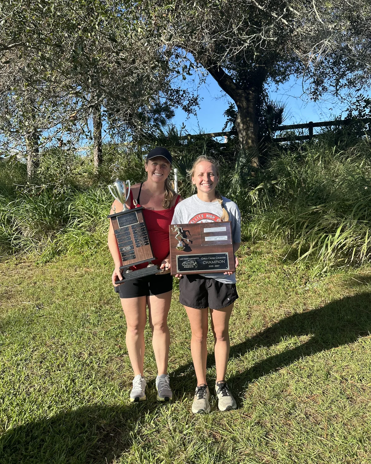 Satellite coaches Brianna Basford and Chelsea Basford Cone standing under a tree at Wickham Park, one holding the Cape Coast Conference trophy and the other the girls cross country champion plaque