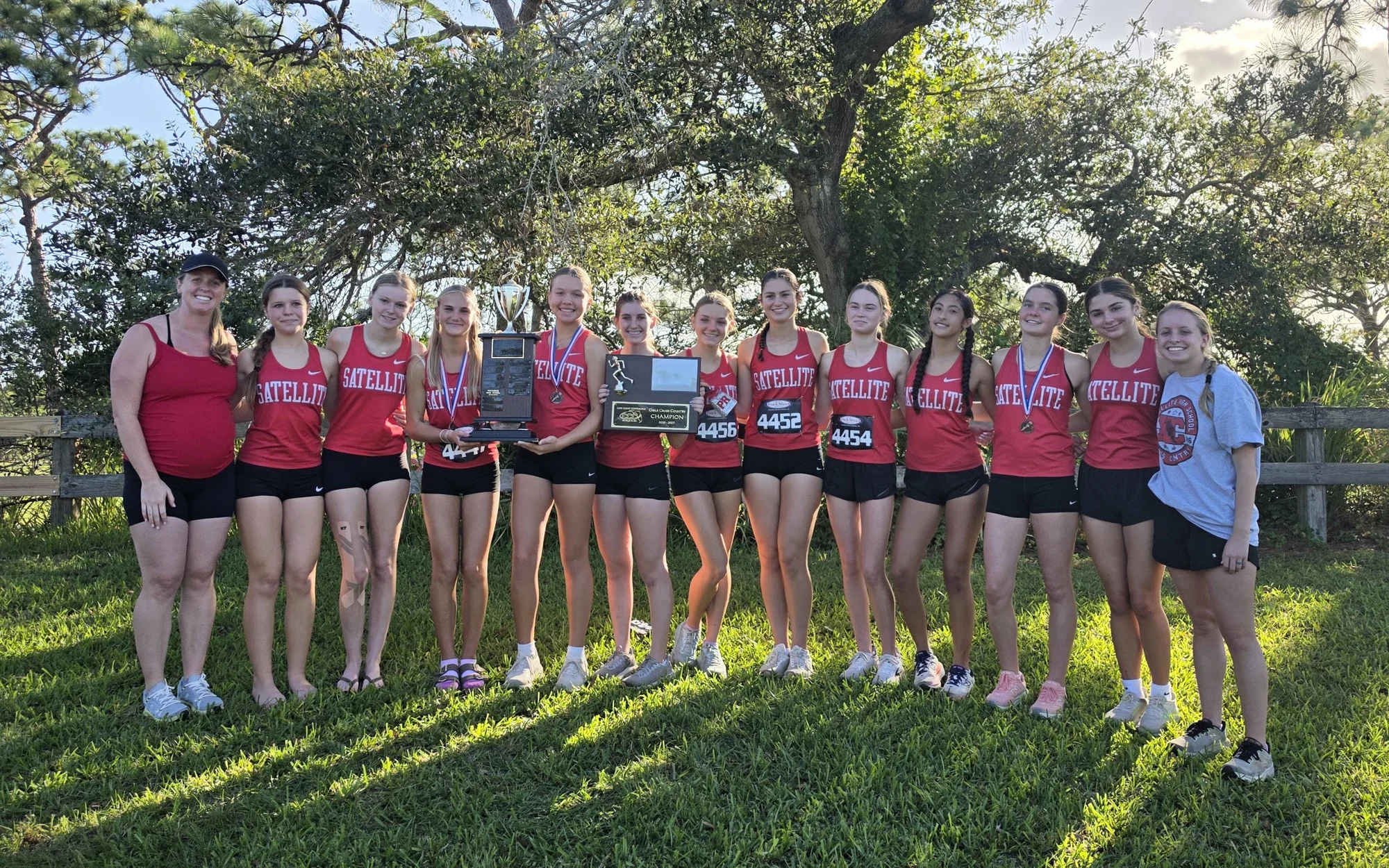 The Satellite girls cross country team in red Satellite singlets, holding the Cape Coast Conference trophy and champion's plaque under a live oak at Wickham Park, with their two coaches at either end