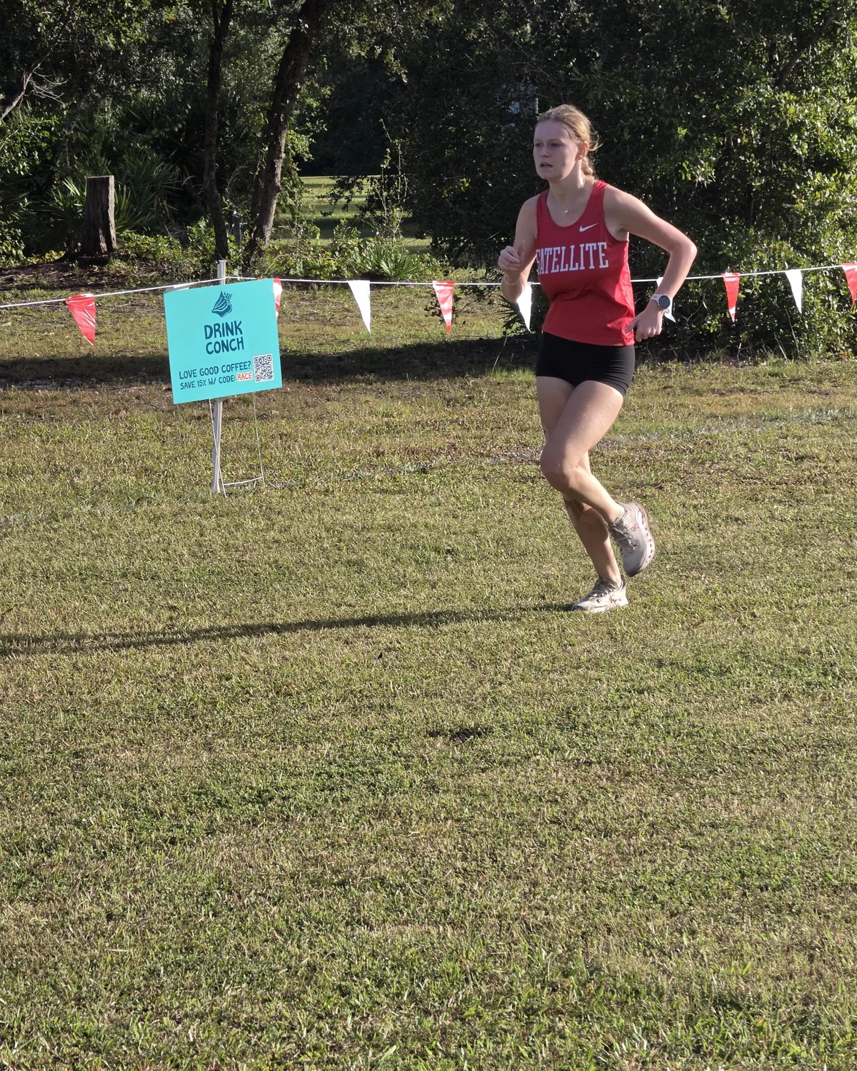 A Satellite runner in a red Satellite singlet and black shorts running along the course tape at Wickham Park, past a sponsor sign