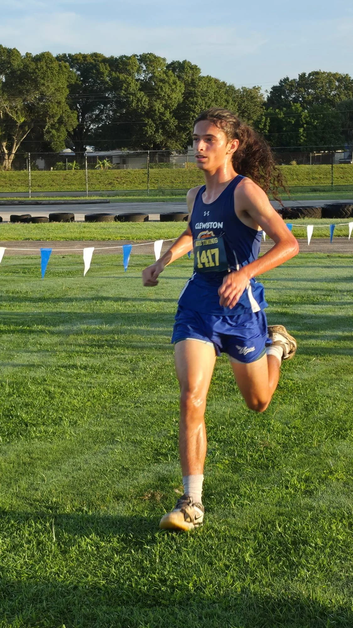 A Clewiston runner with long hair, bib 1417, running on grass beside a line of pennants