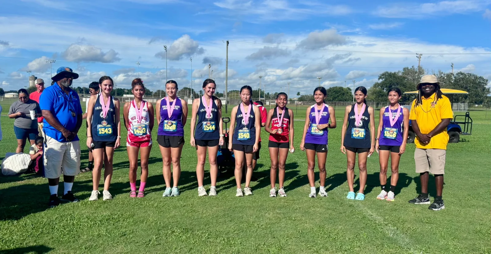 The girls' medalists at the Clewiston XC Sugar Run Invite in a line with pink-ribbon medals