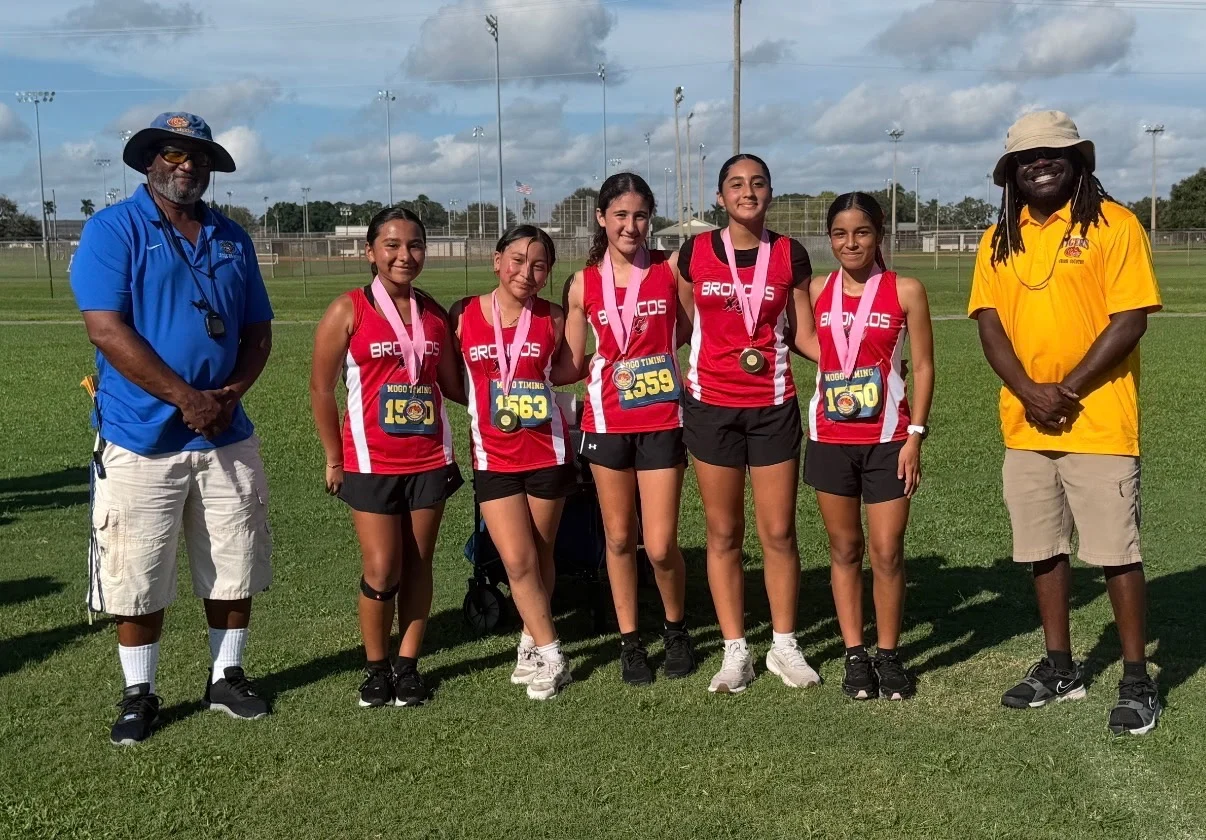 Five LaBelle Middle School girls in red Broncos singlets with medals, between two meet officials