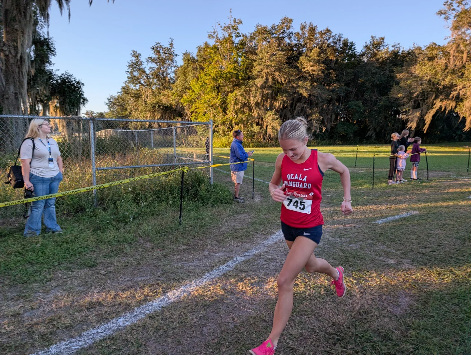Annalise Hallick of Vanguard, bib 745, in a red Ocala Vanguard singlet and pink shoes, running past a spectator and a chain-link fence