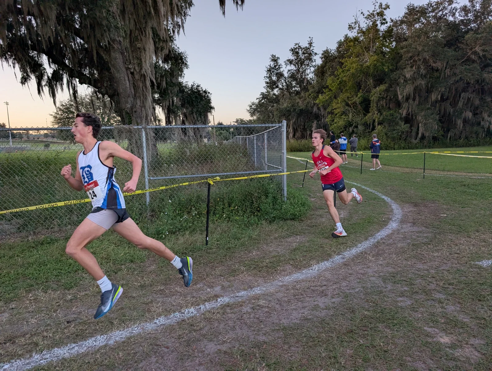 Asher Inman of Belleview, bib 24, in a white and blue Belleview singlet, rounding the fenced curve with a Vanguard runner behind him