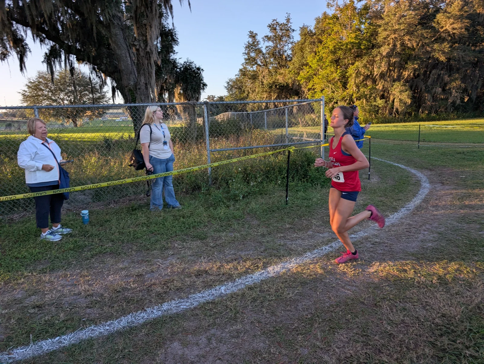 Lyla Hallick of Vanguard, bib 746, in a red Ocala Vanguard singlet, running alone on the fenced curve with spectators behind her