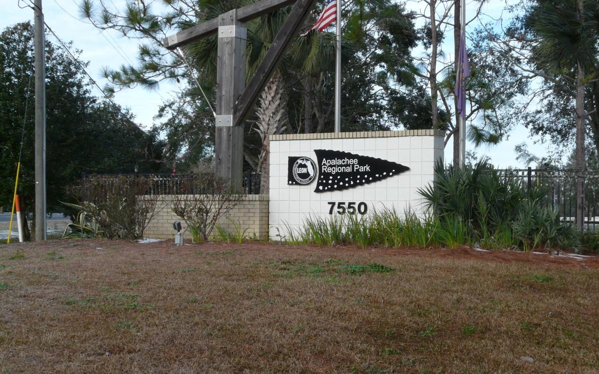 The Apalachee Regional Park entrance sign, with the park name on a black arrowhead and the street number 7550, under pines on Apalachee Parkway in Tallahassee